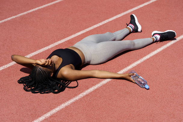Black female beginner feels powerless after running long distance on hot summer day. Exhausted African American woman lies on red track holding bottle of water and covering eyes with hand from sun - Zdjęcie, obraz