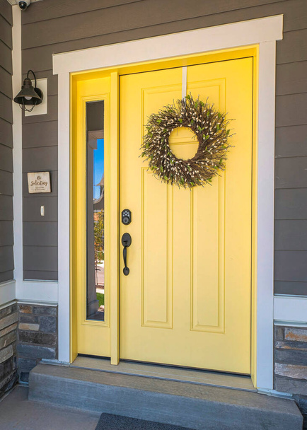 Vertical Yellow front door with wreath and sidelight at the entrance of a house in Utah. Home exterior with gray wood vinyl and stone veneer sidings and a porch with wall lamp near the window. - Foto, Imagem