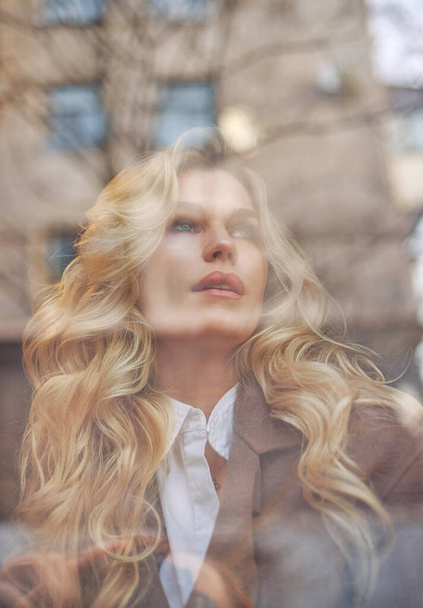 Beautiful elegant woman in a brown coat sits thoughtfully behind the window, glass in the reflections of the street, autumn mood - Fotografie, Obrázek
