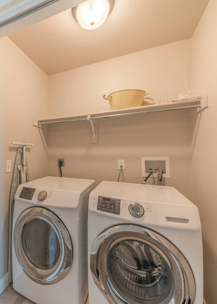 Vertical Reached in laundry with white bi-fold doors and laundry units below the shelf. Interior of a house with laundry room near the stairs and room with window and carpeted flooring. - Photo, image