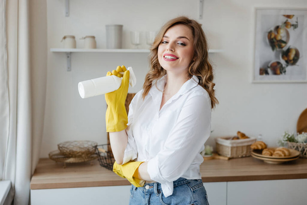 A girl in yellow gloves holds a cleaning spray on the background of the kitchen. High quality photo - Foto, imagen