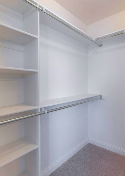 Vertical Small white walk-in closet with brown carpet flooring. There is a shelving unit with metal rods on the left and hooks on the right. - Foto, Imagem