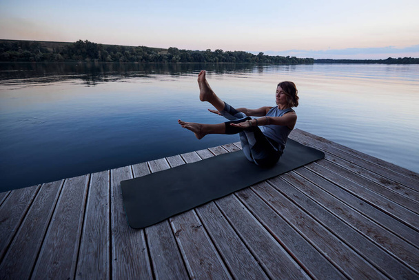 A fit yogi woman is practicing yoga on a dock at dusk. She is in the Boat yoga posture. - Foto, imagen