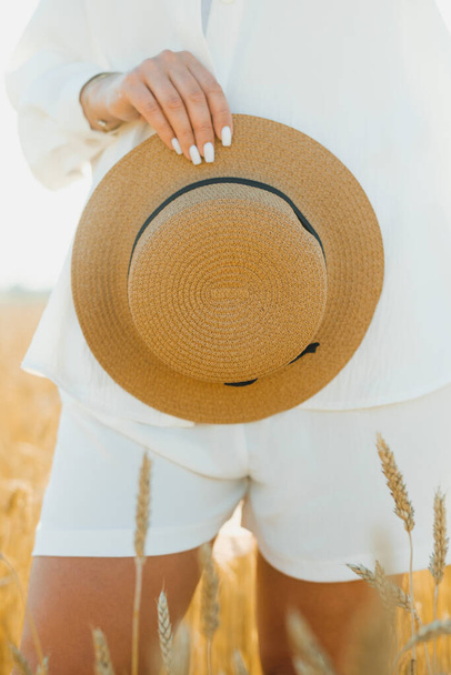 Κοντινό πλάνο με γυναικείο χέρι να κρατάει ψάθινο καπέλο σε χωράφι με σιτάρι μια ηλιόλουστη καλοκαιρινή μέρα. Η έννοια της υπαίθριας αναψυχής, ένα ταξίδι στο χωριό - Φωτογραφία, εικόνα