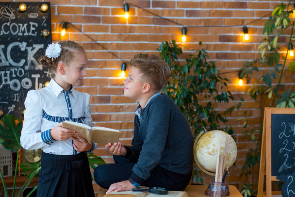 Schoolchildren - a boy and a girl are reading a book in the classroom against the backdrop of a luminous garland, fresh flowers and a globe with a magnifying glass - Photo, Image