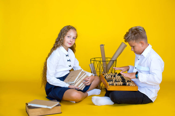 The fair-haired girl and boy - schoolchildren are sitting on a yellow background. The girl is holding books and the boy is holding wooden abacus. - Foto, Imagem