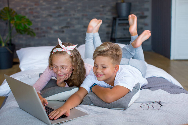 Светловолосые дети - мальчик и девочка учатся и играют на ноутбуке, лежащем на кровати - Фото, изображение