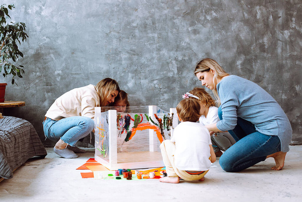 Attentive little girls sitting on haunches on art lesson, painting with women mothers teachers with brushes on stretch film wrapping around turned table legs in grey room. Early development, pairwork. - Photo, Image