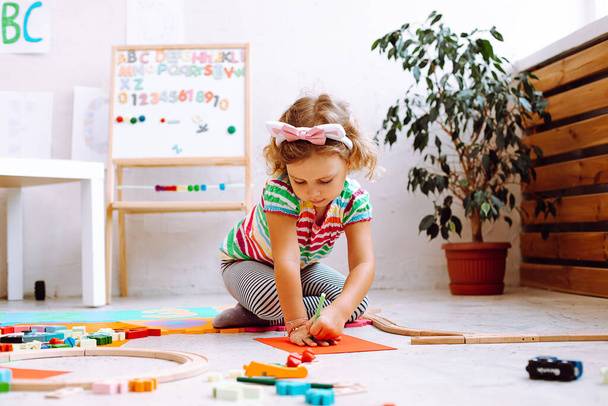 Portrait of cute little girl with short fair hair wearing T-shirt, trousers, sitting near magnetic board with numbers, alphabet, drawing outline of hand on red sheet of paper around different toys. - Foto, Bild