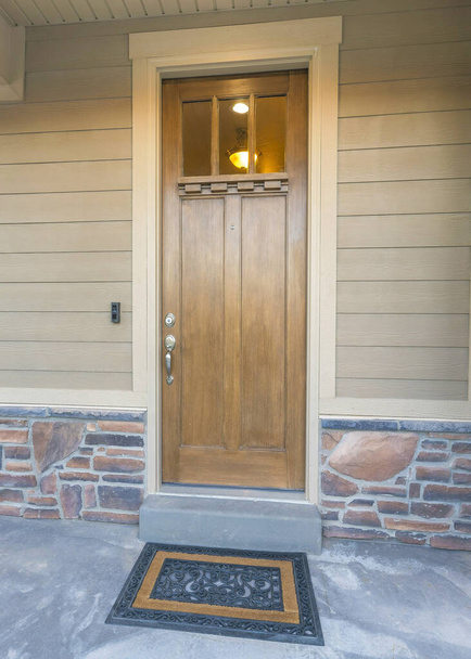 Vertical Wooden front door with glass panes and stylish doormat. Front porch of a house with railings and an exterior of beige vinyl wood and stone veneer sidings. - Photo, Image