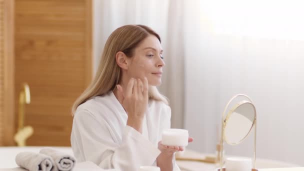 Anti aging beauty care. Attractive middle aged caucasian lady applying skin lifting cream on her face, looking at mirror at home in morning, slow motion, free space - Video, Çekim