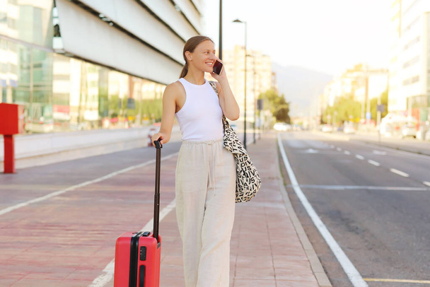 young beautiful woman with red suitcase calling smartphone while waiting for a taxi cab in the city. stylish smiling girl tourist on summer holiday vacation. new trip. copy space. - Foto, Imagen