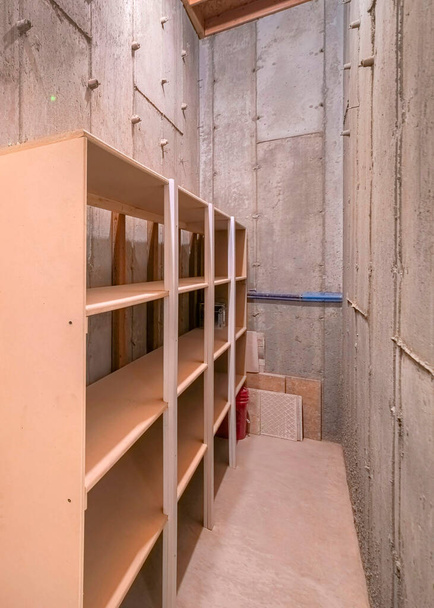 Vertikaler unvollendeter schmaler Lagerraum mit Betonwänden und -böden. Innenraum eines fensterlosen Raumes mit einer Reihe leerer Regale an den Betonwänden. - Foto, Bild