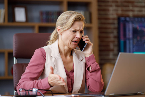 Serious businesswoman having unpleasant phone conversation working on laptop sitting at office. Female trader listening bad news, feels desperate, receive bad information about monetary investments. - Foto, afbeelding