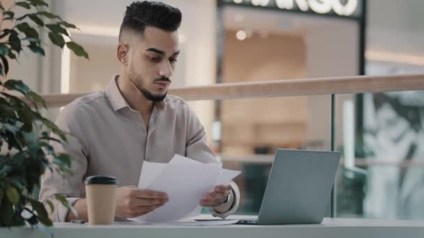 Serious busy multitasking Arabian Indian businessman checking corporate paperwork sitting at office cafe desk. Male worried entrepreneur working with documents analyzing financial papers with laptop - Materiał filmowy, wideo