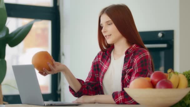Καυκάσια νεαρή γυναίκα blogger τροφίμων κοκκινομάλλα διατροφολόγος κάθεται στην κουζίνα δείχνει σε κάμερα γκρέιπφρουτ εσπεριδοειδών συνιστά online υπηρεσία των προϊόντων παράδοσης φρέσκα φρούτα χρησιμοποιεί φορητό υπολογιστή τηλεδιάσκεψη κλήσης - Πλάνα, βίντεο