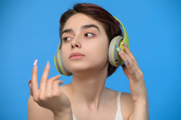 Portrait of cute young bare shoulders brunette with extravagant nail art listening to music using bluetooth wireless headphones, studio shot - Foto, immagini