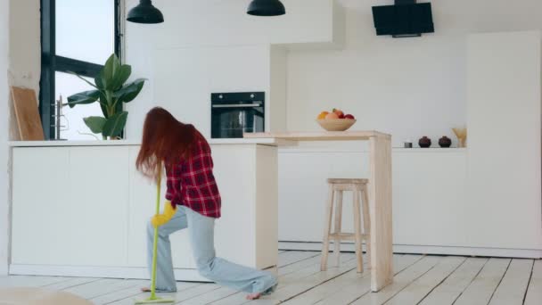 Active happy caucasian housewife amusing young redhead woman with long hair energetic dancer moving to music cleaning apartment flat washing floor with mop dancing in living room housekeeping chores - Imágenes, Vídeo