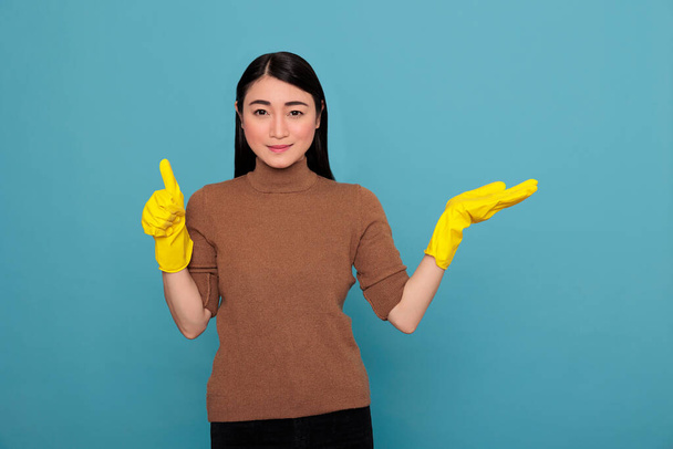 χαμογελαστή χαρούμενη νεαρή Ασιάτισσα από τις δουλειές που δείχνουν το χέρι στην άκρη στο χώρο των αντιγράφων και φορώντας κίτρινο γάντι, Καθαρισμός έννοια σπίτι, αισιόδοξη ικανοποιημένη θετική γυναικεία αντίχειρες επάνω - Φωτογραφία, εικόνα