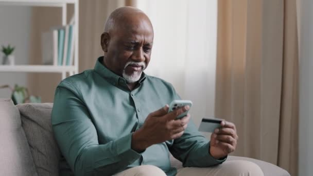 Confused puzzled senior African old bald senior elderly businessman with gray beard has problem with online payment upset retired man error in financial app smartphone money problem transfer at home - Felvétel, videó