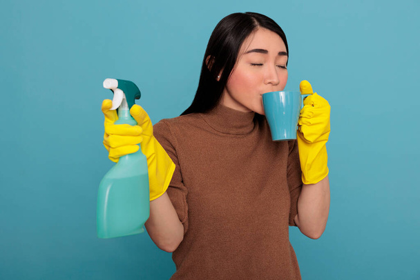 Tired exhausted young asian housewife taking coffee during work break while holding a sponge in the other hand, Cleaning home concept, Refreshment tea take some rest from day to day chores, - 写真・画像