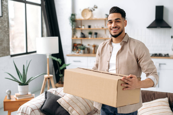 Happy confident indian or arabian guy, stand at home in living room, holding a large cardboard box, received a long-awaited parcel from the online store, preparing to unpack, looks at camera, smiles - Photo, image