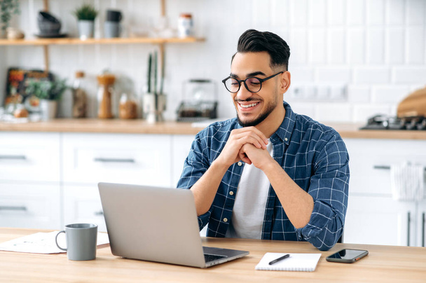 Photo of attractive positive indian or arabian guy with glasses, student or freelancer, working remotely, sitting at home in the kitchen with laptop, looking at the screen, thinking, smile happily - Photo, image