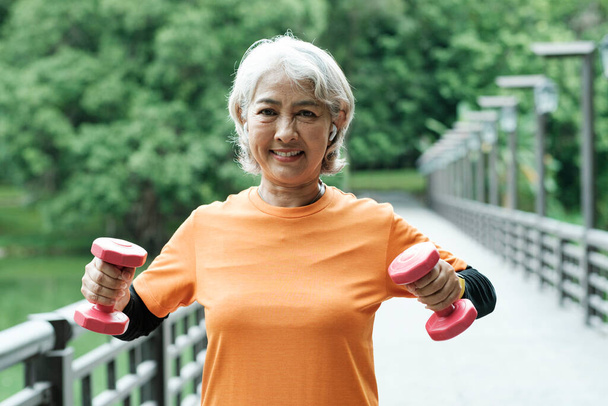 Athletic Senior woman lifts dumbbells while doing fitness before running in park, health concept. - Foto, imagen