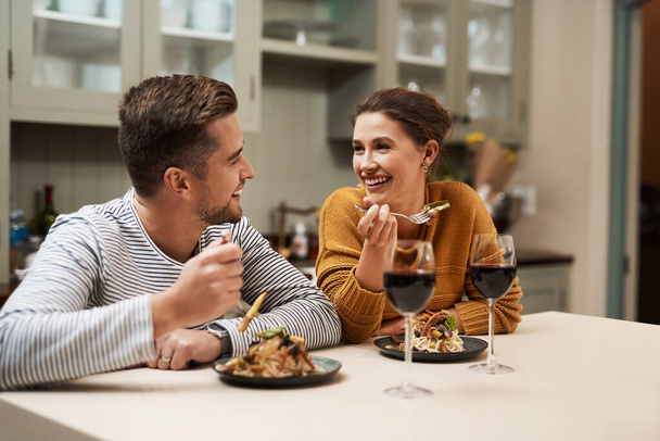 Who doesnt love a home cooked meal. an affectionate young couple smiling at each other while enjoying dinner in their kitchen at home - Фото, изображение