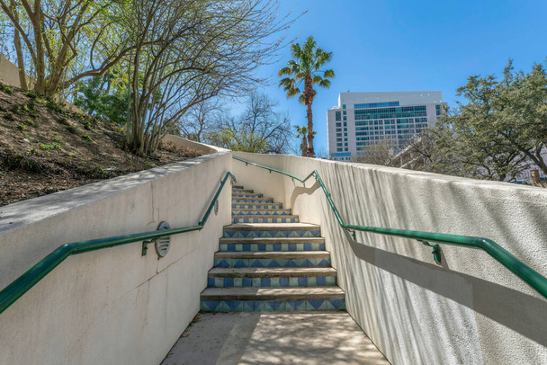 Staircase with concrete half wall and tile risers at San Antonio, Texas. There is a wall-mounted metal handrail and a slope on the left with trees and view of buildings at the back against the sky. - Photo, image