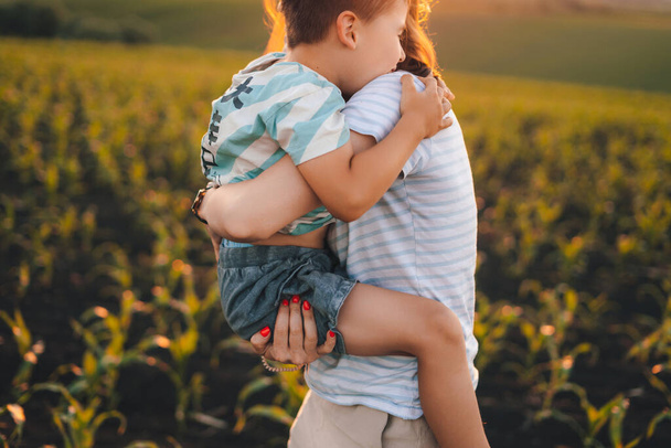 Πορτρέτο ενός μικρού αγοριού που στέκεται στην αγκαλιά της μαμάς που περνάει μέσα από το χωράφι με τα καλαμπόκια. Η μητέρα περπατάει με ένα παιδί σε ένα χωράφι με σιτάρι. Χαρούμενα οικογενειακά ταξίδια. - Φωτογραφία, εικόνα