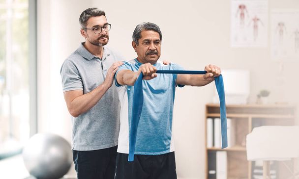 Growing stronger with every stretch. a senior man exercising with a resistance band during a rehabilitation session with his physiotherapist - Valokuva, kuva