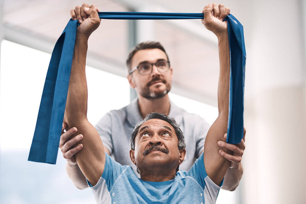 Were getting fitter by the day together. a senior man exercising with a resistance band during a rehabilitation session with his physiotherapist - Foto, Bild