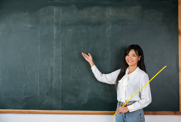 Back to school concept. Happy beautiful young woman standing hold pointer to back board, Asian female teacher smiling with wooden stick pointing to blackboard at school in classroom, Education - Photo, image