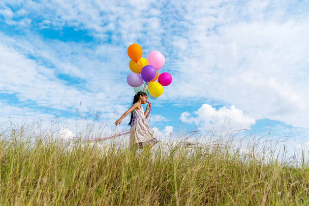 Χαρούμενο χαριτωμένο κορίτσι κρατώντας μπαλόνια που τρέχουν στο πράσινο λιβάδι λευκό σύννεφο και μπλε ουρανό με την ευτυχία. Χέρια κρατώντας ζωντανή μπαλόνια αέρα παίζουν στο πάρτι γενεθλίων χαρούμενες στιγμές καλοκαίρι στο φως του ήλιου εξωτερική - Φωτογραφία, εικόνα