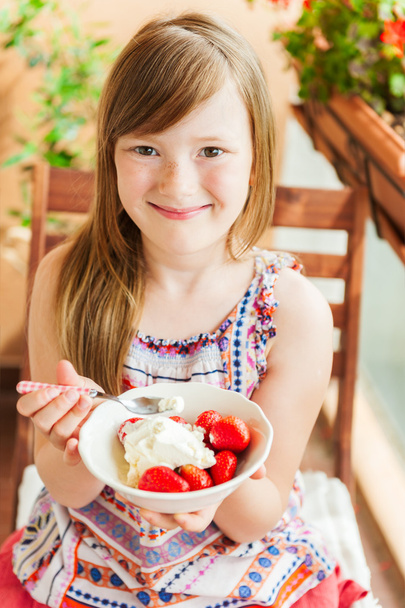 Χαριτωμένο κοριτσάκι που τρώει φράουλα με παγωτό σε ένα μπαλκόνι, μια ωραία ηλιόλουστη ημέρα - Φωτογραφία, εικόνα