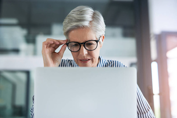 I cant believe what Im seeing right now. an attractive mature businesswoman adjusting her glasses while working on a laptop in her office - Foto, Imagen