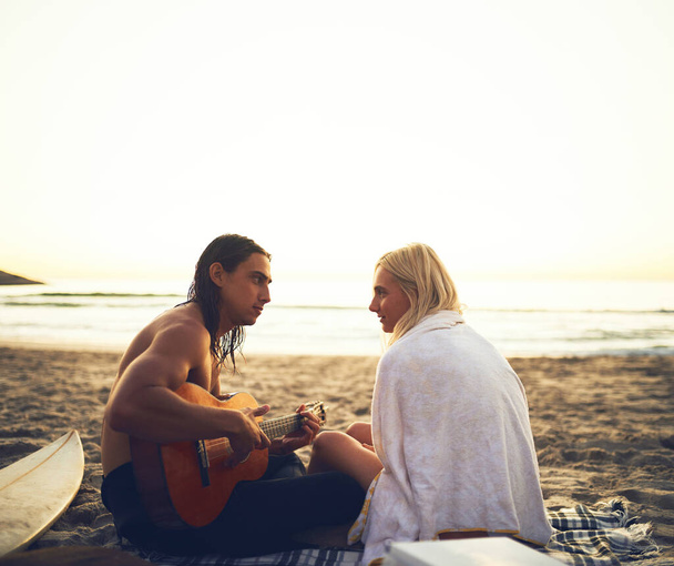 Το ξέρω αυτό το τραγούδι. Στιγμιότυπο ενός νεαρού στοργικού ζευγαριού να κάνει καντάδα ο ένας στον άλλο κατά τη διάρκεια ενός ραντεβού στην παραλία το ηλιοβασίλεμα - Φωτογραφία, εικόνα