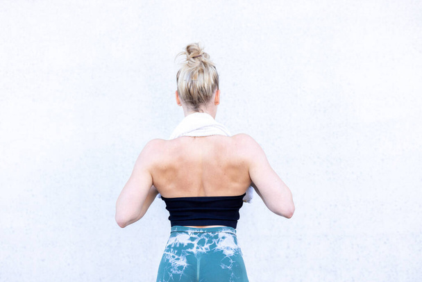 Half Body Rear View Shot of a Gym Fit Caucasian sporty woman dressed in sportswear and with her hair up holding a towl around her neck with her hands and standing in the gym. In background wall, backs - Photo, Image