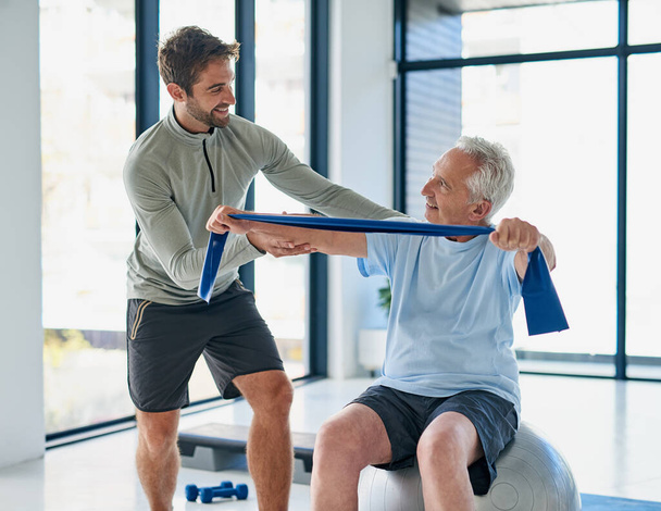 This routine has been good for me. a young male physiotherapist helping a senior man stretch with a stretching band in a fitness centre - Foto, Bild