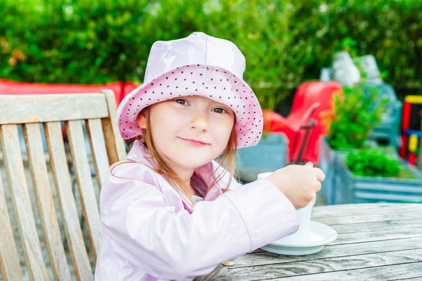 Linda niña bebiendo chocolate caliente en un café, usando, abrigo de lluvia rosa y sombrero
 - Foto, Imagen
