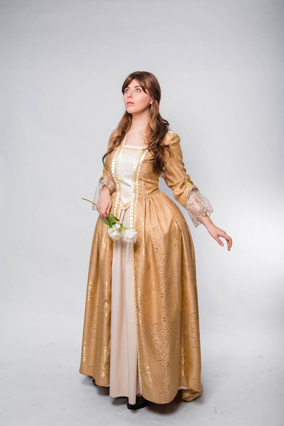 Full length portrait of a woman in a gold dress in the style of the rococo era, standing and posing with a flower in hand isolated on a white background. - Zdjęcie, obraz