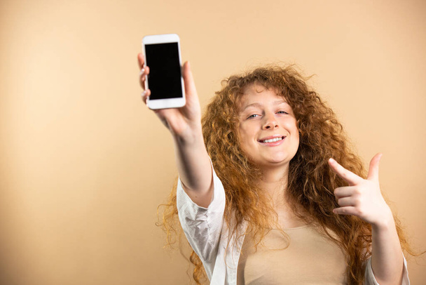 Portrait of cool pretty young redhead woman holding a smartphone in raised forward hand with black screen over golden background. High quality photo - Foto, Imagen