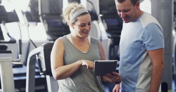 Personal trainer or coach talking to a client about planning his exercise schedule on a tablet. A happy female fitness instructor consulting a fit man about this workout or performance at the gym. - Felvétel, videó