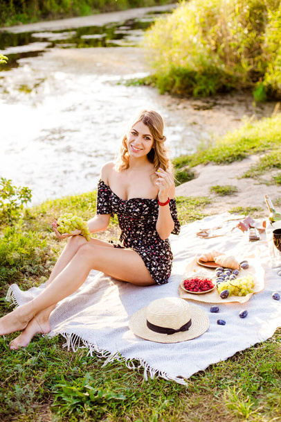 Young attractive girl with dark hair in a summer dress on a picnic in nature by the river in summer at sunset. Healthy life. - Foto, Imagen