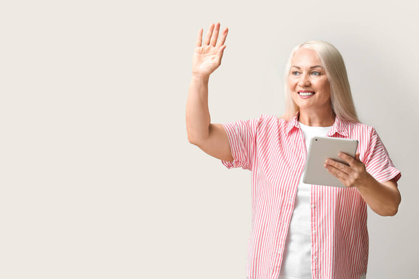 Mature woman with tablet computer for navigation waving hand on light background - Foto, Imagem