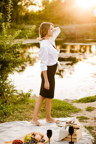 Young attractive girl with dark hair in a white shirt on a picnic in nature by the river in summer at sunset. Healthy life. - Φωτογραφία, εικόνα
