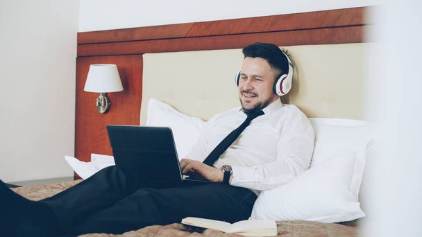 Happy cheerful businessman in headphones working at laptop computer and listening music smiling while lying in bed at hotel room. Travel, business and people concept - Фото, зображення