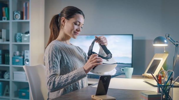 Happy young woman sitting at desk at home and holding a VR headset, virtual reality and technology concept - Φωτογραφία, εικόνα