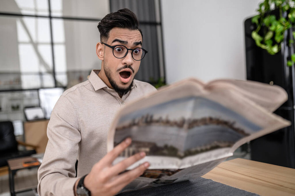 Shock. Young adult attractive man in glasses with open mouth surprised looking at newspaper sitting at table indoors - 写真・画像